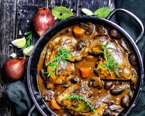 Herzhaftes Coq au Vin in einem gusseisernen Topf, mit Pilzen, Karotten und Kräutern garniert. Daneben Zwiebeln und Knoblauch.