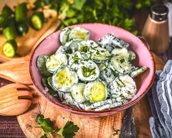 Omas Gurkensalat mit Joghurt, frisch zubereitet und mit Kräutern verfeinert, serviert in einer rosa Schüssel auf einem Holzbrett.