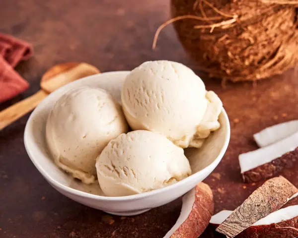Schaufeln cremiges Kokoseis, perfekt zum Kokoseis selber machen mit Eismaschine. Daneben Kokosnussstücke.