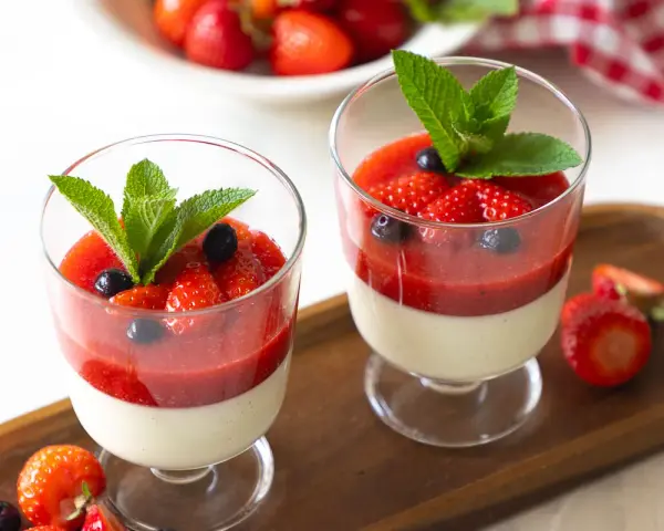 Zwei Gläser mit cremiger Panna cotta, garniert mit Erdbeersauce, frischen Erdbeeren, Blaubeeren und Minze.