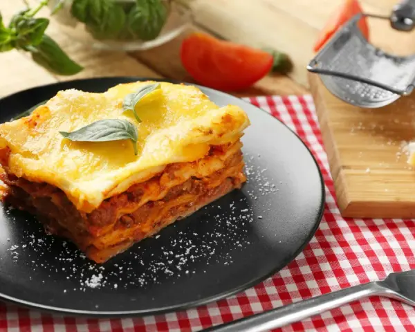 Ein Stück Lasagne mit Hackfleischsauce und cremiger Bechamelsoße, garniert mit Basilikum. Daneben ein Stück Parmesan und eine Gabel.