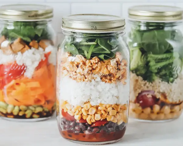 Drei Gläser mit geschichtetem Salat: Reis, Mais, Bohnen, Tomaten, Karotten und Spinat. Perfekt für unterwegs!