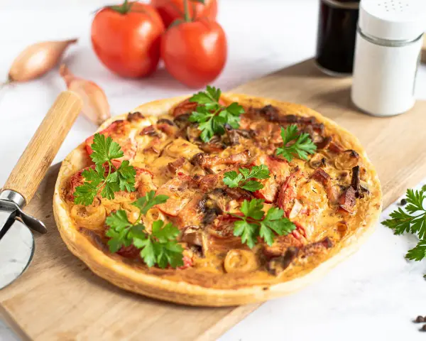 Flammkuchen mit Pfifferlingen, garniert mit Petersilie, auf einem Holzbrett. Tomaten und Schalotten im Hintergrund.