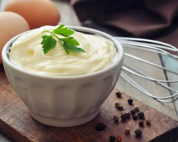 Cremige Mayonnaise in einer Schüssel, garniert mit Petersilie. Eier und ein Schneebesen deuten auf ein hausgemachtes mayonnaise rezept hin.