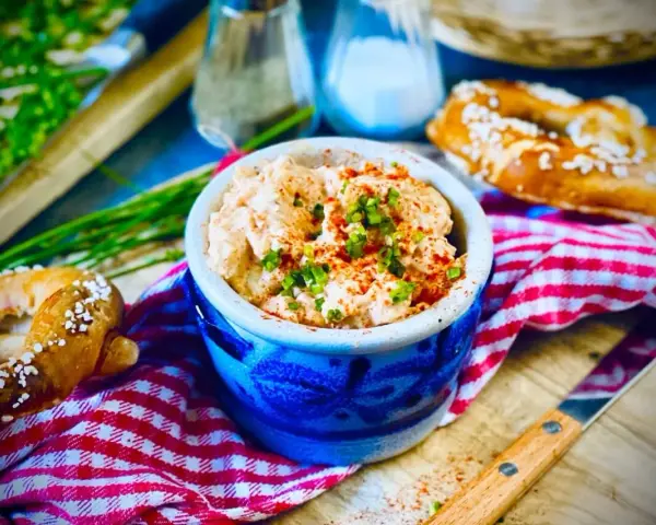 Leckerer Obazda im blauen Steingut-Töpfchen, bestreut mit Schnittlauch und Paprikapulver. Dazu Brezeln und ein Messer. Perfekt für ein Obazda Rezept!