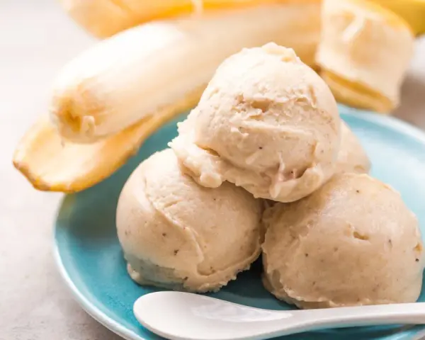 Cremiges Bananeneis, ganz einfach eis selber machen ohne eismaschine. Dazu reife Bananen und eine weiße Löffel auf blauem Teller.