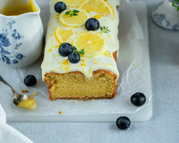 Ein saftiger zitronen schmand kuchen, dekoriert mit Zitronenscheiben, Blaubeeren und Zitrusschale. Daneben eine Kanne mit Zitronensauce.