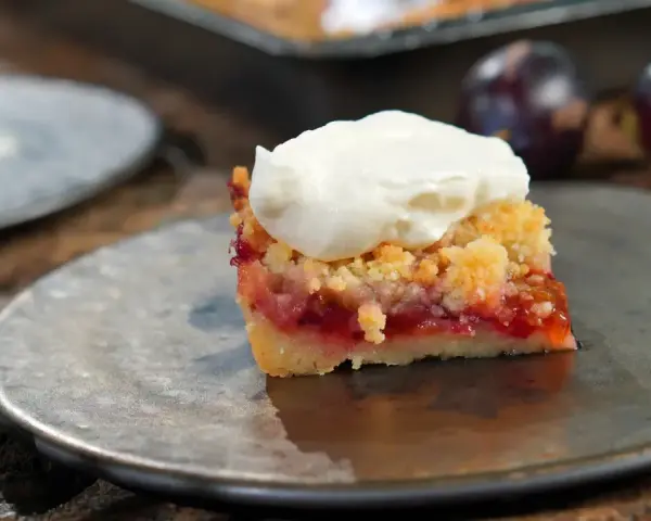 Ein Stück Zwetschgenkuchen mit Streuseln und Sahne auf einem Teller, daneben frische Zwetschgen.