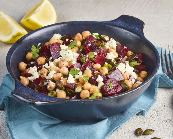 Ein bunter rote Bete Kichererbsen Salat mit Feta und Kürbiskernen in einer blauen Schüssel, garniert mit Petersilie und Zitronenspalten.