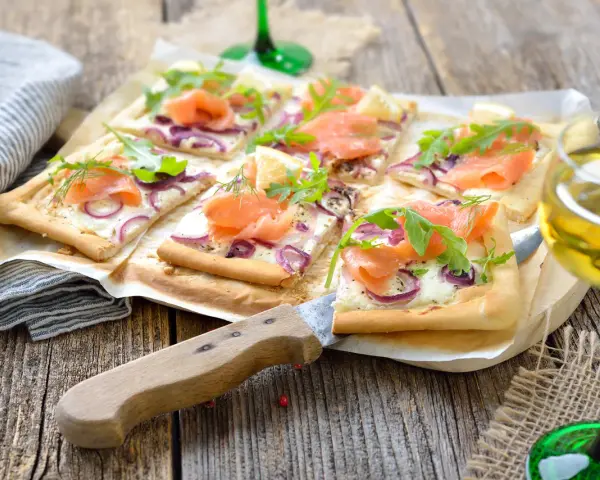Flammkuchen mit Lachs und Crème fraîche, garniert mit Rucola und Dill, auf rustikalem Holztisch.
