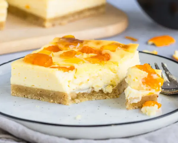 Ein Stück Mandarinen Schmand Kuchen Blech mit fruchtiger Glasur auf einem Teller, daneben ein Stück mit einer Gabel.