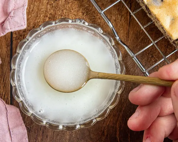 Ein Löffel mit zuckerguss ohne puderzucker in einer Schale. Daneben ein Stück Kuchen auf einem Gitter.