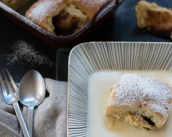 Buchteln original rezept: Ein Buchtel in Vanillesauce, bestäubt mit Puderzucker. Im Hintergrund weitere Buchteln im Ofen.