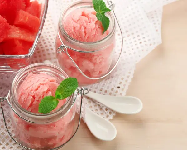 Erfrischendes Meloneneis selber machen: Rosa Sorbet in Gläsern mit Minze, daneben eine Schale mit Wassermelonenwürfeln.