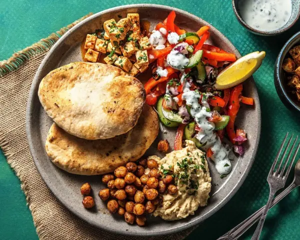 Ein bunter griechischer Salat mit Hummus, gerösteten Kichererbsen und Pitabrot. Dazu gewürfelter Tofu und ein Zitronenschnitz.
