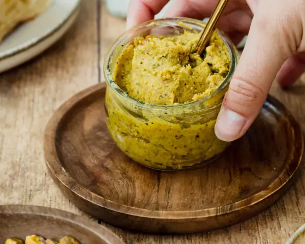 Ein Glas mit selbstgemachter Pistaziencreme, die mit einem goldenen Löffel umgerührt wird. Perfekt zum Brot oder als Dip.