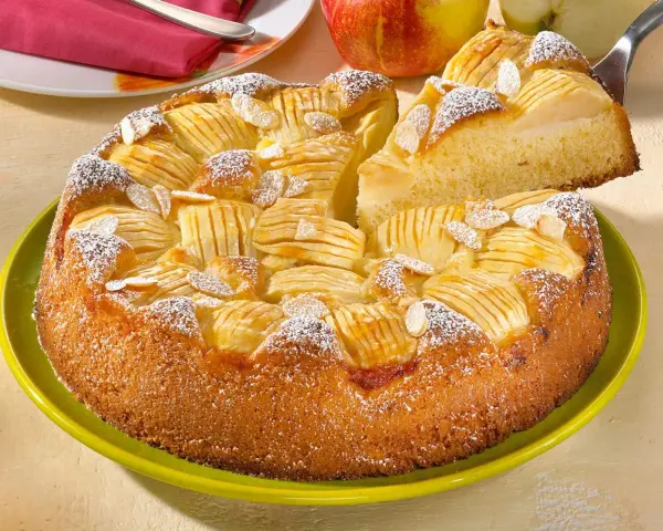 Ein saftiger, versunkener Apfelkuchen mit Mandelsplittern und Puderzucker bestreut, wird auf einem grünen Teller präsentiert.