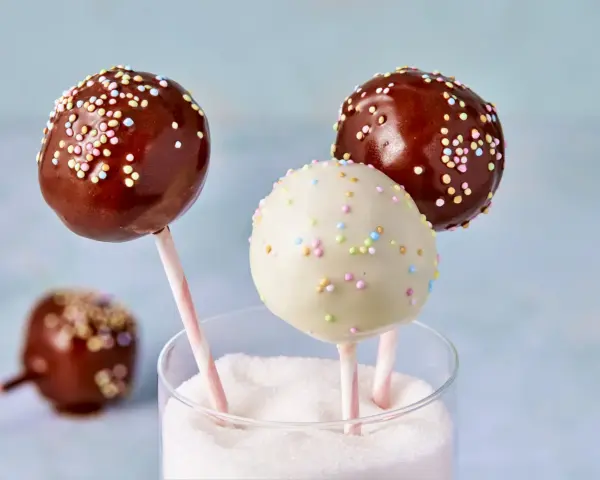 Drei bunte cake pops aus kuchenresten, mit Schokolade und Streuseln verziert, stecken in einem Glas voller Zucker.