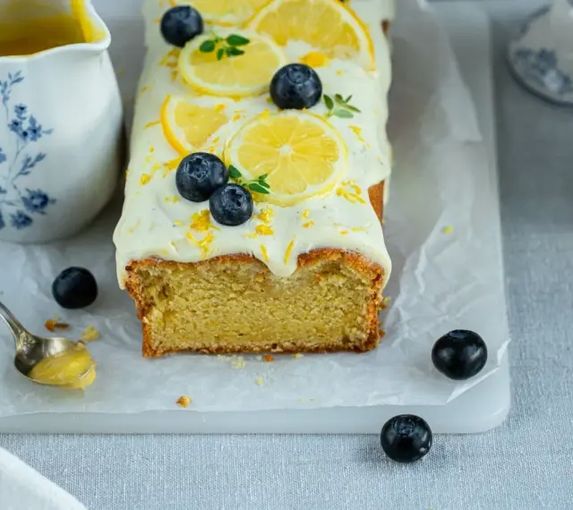 Ein saftiger zitronen schmand kuchen, dekoriert mit Zitronenscheiben, Blaubeeren und Zitrusschale. Daneben eine Kanne mit Zitronensauce.