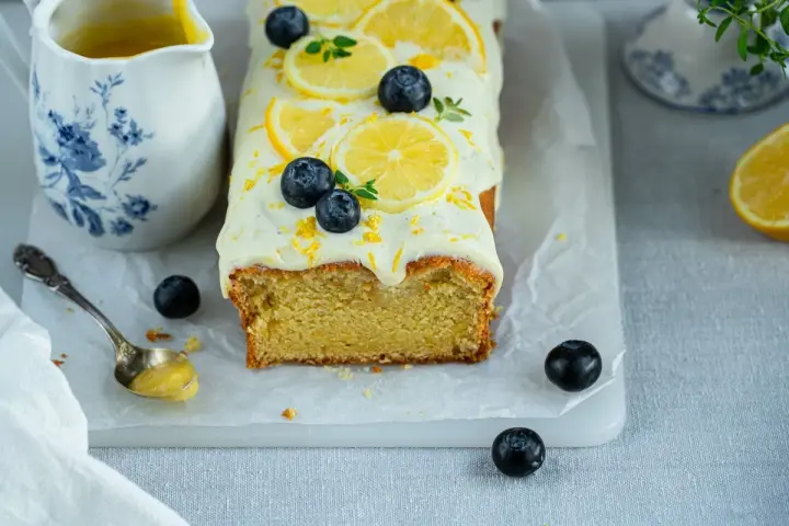Ein saftiger zitronen schmand kuchen, dekoriert mit Zitronenscheiben, Blaubeeren und Zitrusschale. Daneben eine Kanne mit Zitronensauce.