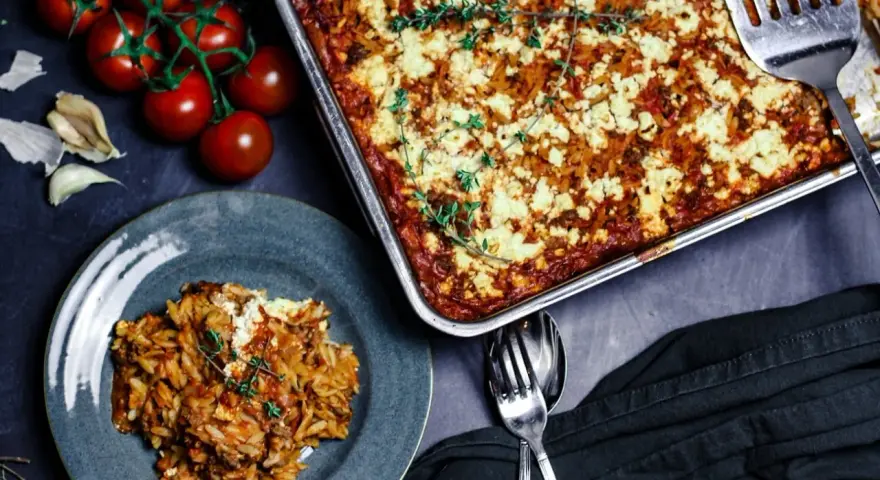 Herzhafter Kritharaki Auflauf mit Hackfleisch und Feta, garniert mit Thymian. Daneben frische Tomaten und Knoblauch.
