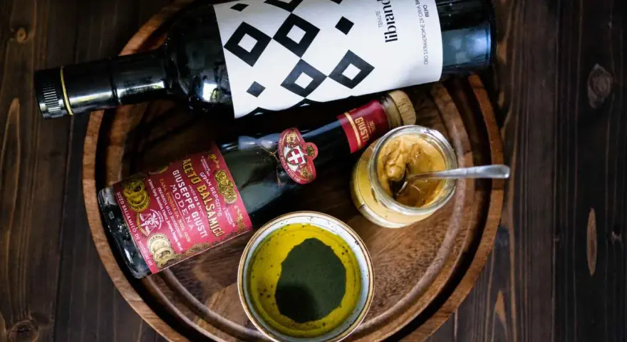 Zutaten für ein köstliches Balsamico-Dressing: eine Flasche Olivenöl, eine Flasche Aceto Balsamico Giuseppe Giusti und eine Schale mit grünen Kräutern.