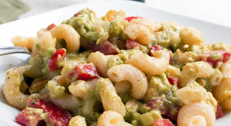 Ein cremiger nudelsalat mit avocado, Tomaten und Gewürzen auf einem weißen Teller.