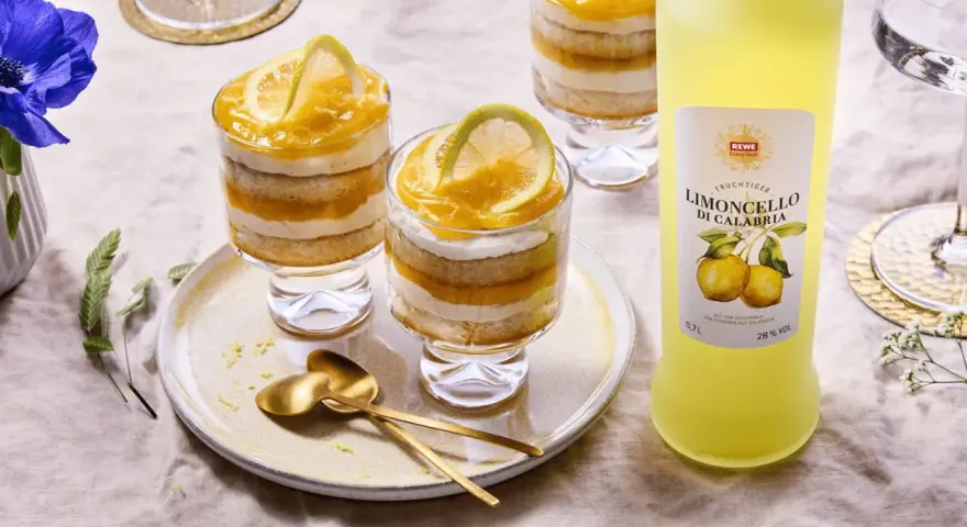 Erfrischendes Limoncello Tiramisu im Glas, geschichtet mit Biskuit und Zitronencreme, garniert mit Zitronenscheiben. Daneben eine Flasche Limoncello.