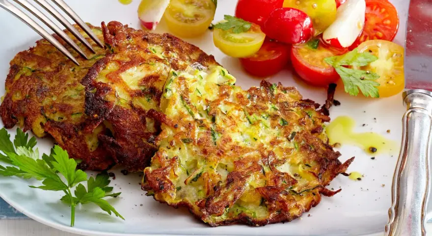 Goldbraune Zucchini-Puffer, knusprig gebraten, serviert mit einem bunten Tomatensalat und frischer Petersilie. Ein Genuss!