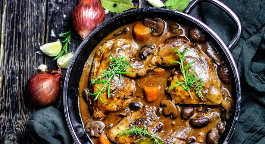 Herzhaftes Coq au Vin in einem gusseisernen Topf, mit Pilzen, Karotten und Kräutern garniert. Daneben Zwiebeln und Knoblauch.