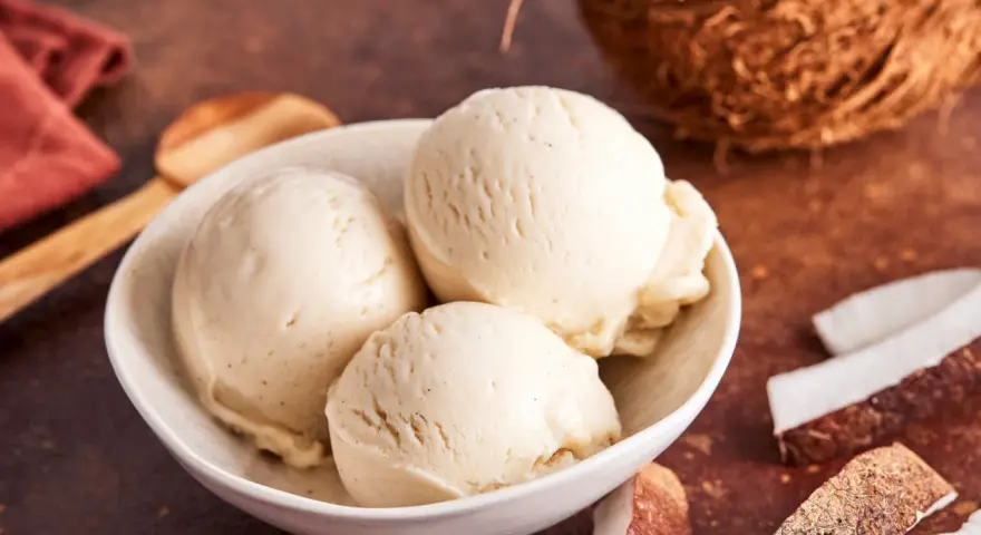 Schaufeln cremiges Kokoseis, perfekt zum Kokoseis selber machen mit Eismaschine. Daneben Kokosnussstücke.