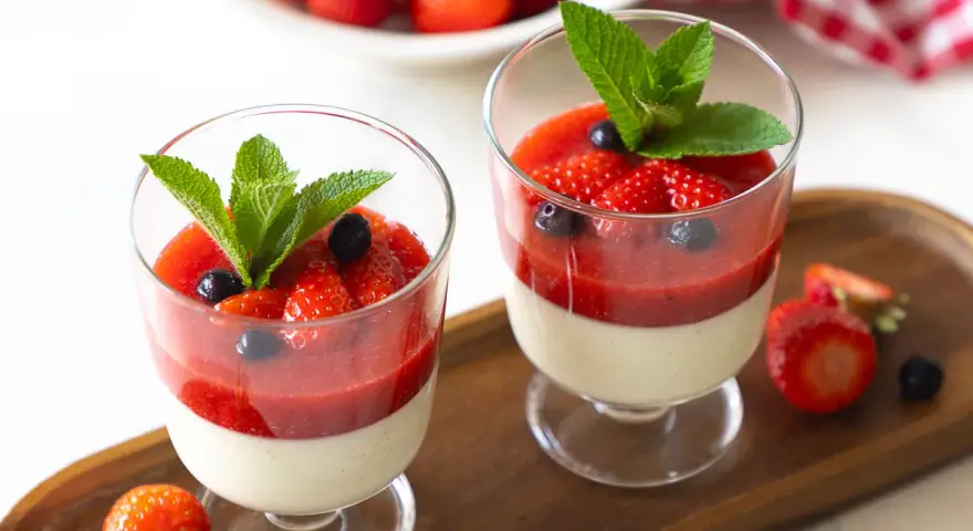 Zwei Gläser mit cremiger Panna cotta, garniert mit Erdbeersauce, frischen Erdbeeren, Blaubeeren und Minze.