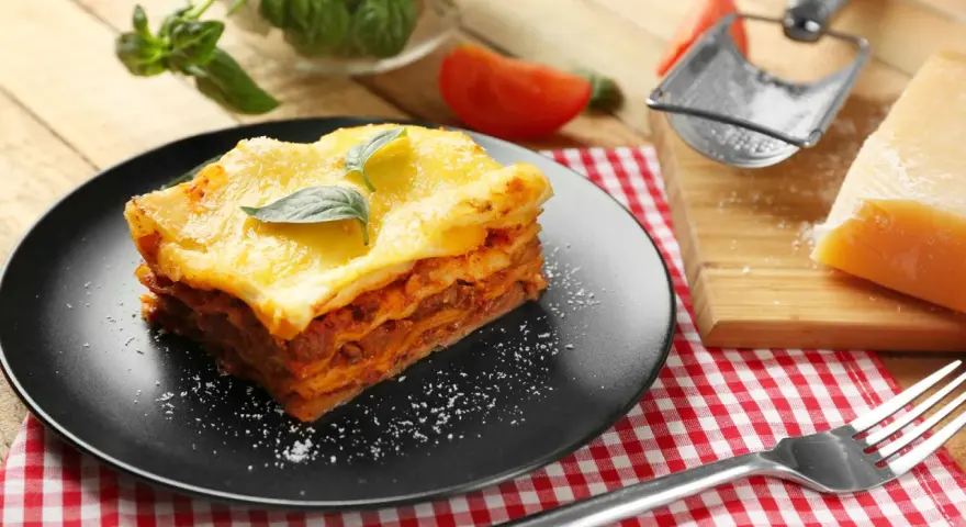 Ein Stück Lasagne mit Hackfleischsauce und cremiger Bechamelsoße, garniert mit Basilikum. Daneben ein Stück Parmesan und eine Gabel.