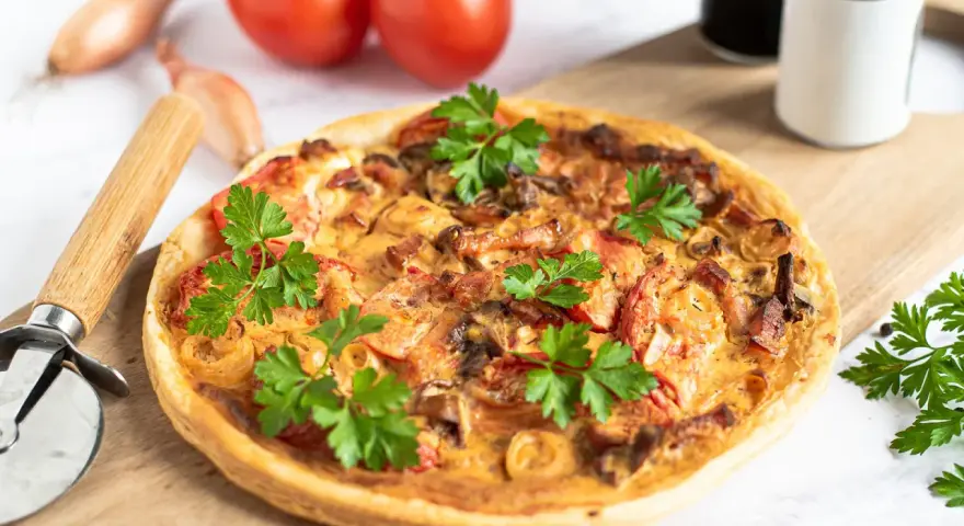 Flammkuchen mit Pfifferlingen, garniert mit Petersilie, auf einem Holzbrett. Tomaten und Schalotten im Hintergrund.