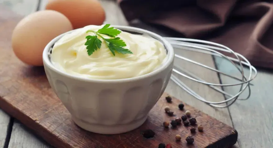 Cremige Mayonnaise in einer Schüssel, garniert mit Petersilie. Eier und ein Schneebesen deuten auf ein hausgemachtes mayonnaise rezept hin.