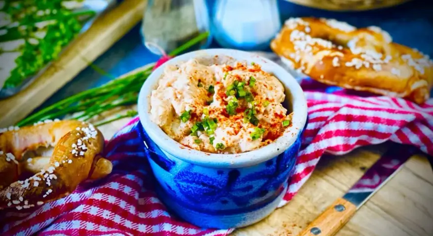 Leckerer Obazda im blauen Steingut-Töpfchen, bestreut mit Schnittlauch und Paprikapulver. Dazu Brezeln und ein Messer. Perfekt für ein Obazda Rezept!
