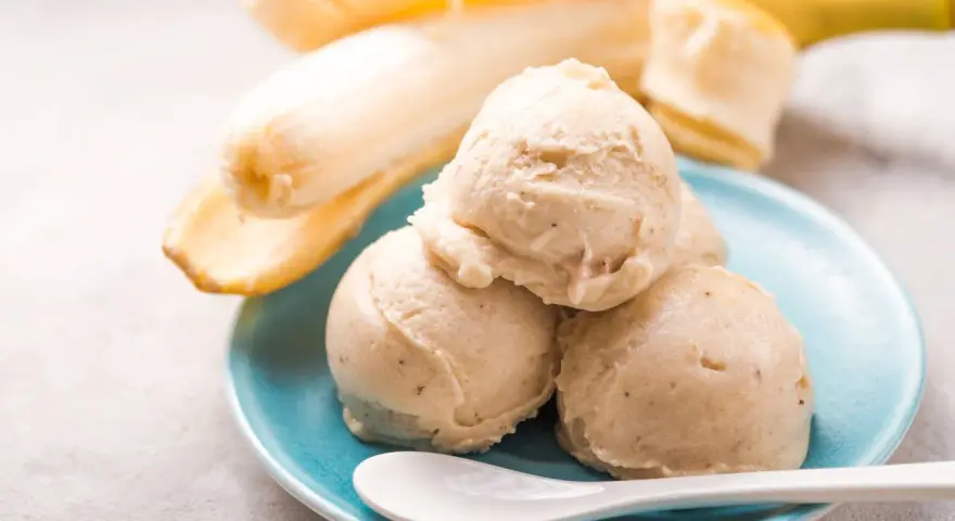 Cremiges Bananeneis, ganz einfach eis selber machen ohne eismaschine. Dazu reife Bananen und eine weiße Löffel auf blauem Teller.