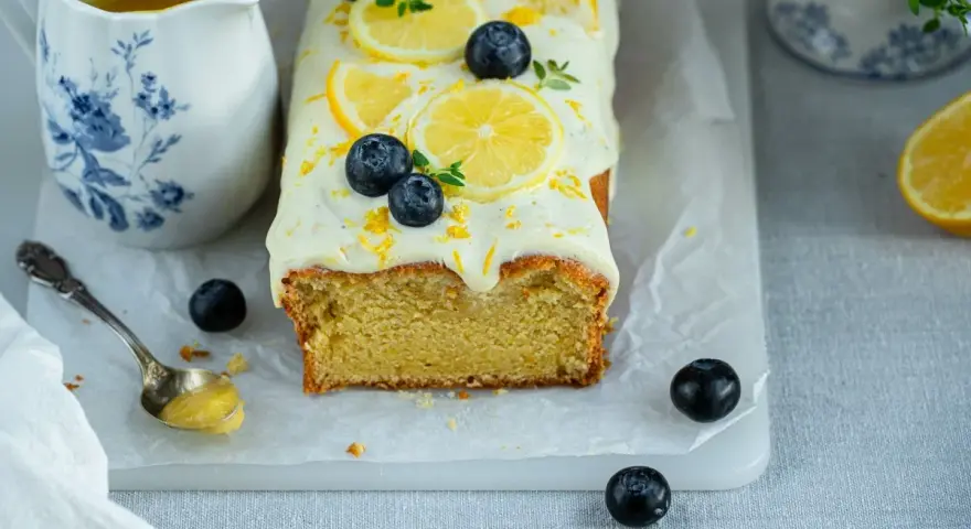 Ein saftiger zitronen schmand kuchen, dekoriert mit Zitronenscheiben, Blaubeeren und Zitrusschale. Daneben eine Kanne mit Zitronensauce.
