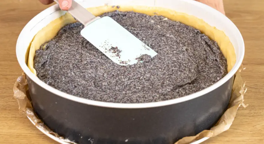 Ein Mohnkuchen mit Puddingfüllung wird in einer Springform vorbereitet. Eine Hand glättet die Mohnmasse mit einem Spatel.