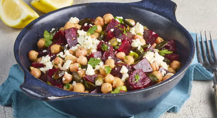 Ein bunter rote Bete Kichererbsen Salat mit Feta und Kürbiskernen in einer blauen Schüssel, garniert mit Petersilie und Zitronenspalten.