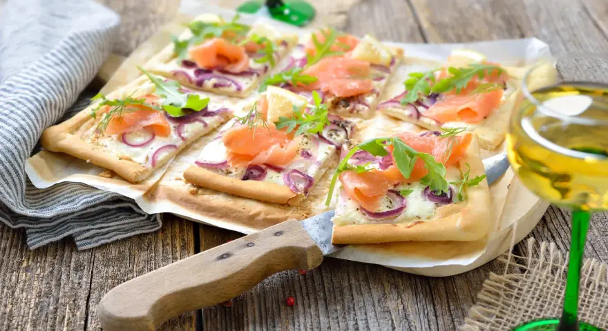 Flammkuchen mit Lachs und Crème fraîche, garniert mit Rucola und Dill, auf rustikalem Holztisch.