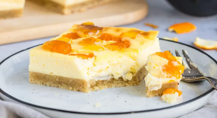 Ein Stück Mandarinen Schmand Kuchen Blech mit fruchtiger Glasur auf einem Teller, daneben ein Stück mit einer Gabel.