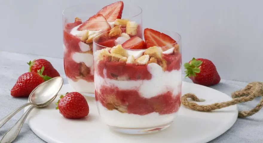 Zwei Gläser mit einem sommerlichen Dessert aus Erdbeeren und Sahne, garniert mit frischen Erdbeeren und Keksen. Ein schnell und einfach zubereitetes Genusserlebnis.