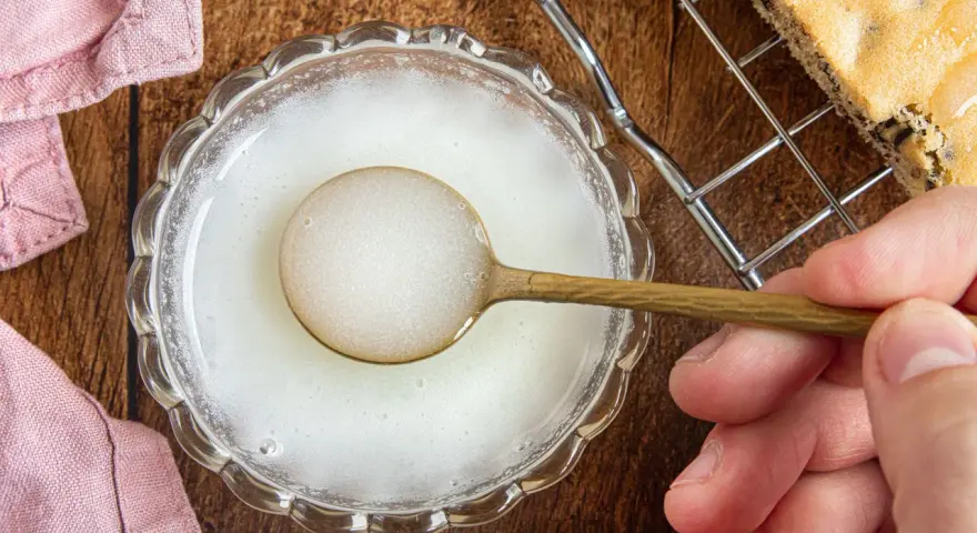 Ein Löffel mit zuckerguss ohne puderzucker in einer Schale. Daneben ein Stück Kuchen auf einem Gitter.