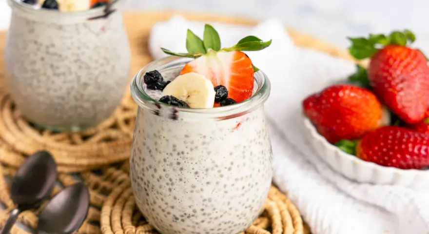 Zwei Gläser mit cremigem Chia Pudding, garniert mit Erdbeeren, Bananen und Blaubeeren. Daneben eine Schale voller frischer Erdbeeren.