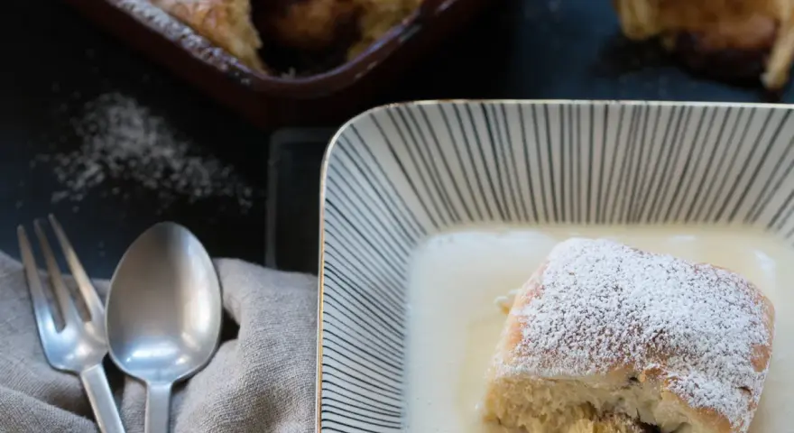 Buchteln original rezept: Ein Buchtel in Vanillesauce, bestäubt mit Puderzucker. Im Hintergrund weitere Buchteln im Ofen.