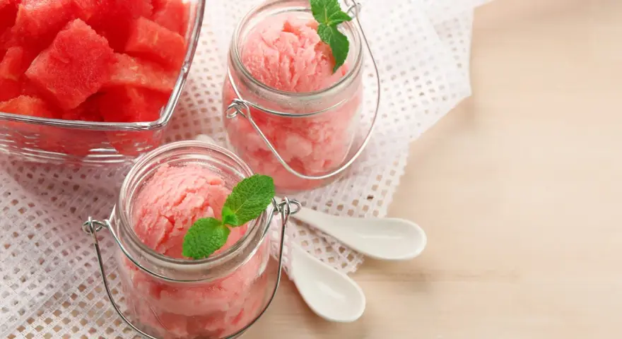 Erfrischendes Meloneneis selber machen: Rosa Sorbet in Gläsern mit Minze, daneben eine Schale mit Wassermelonenwürfeln.