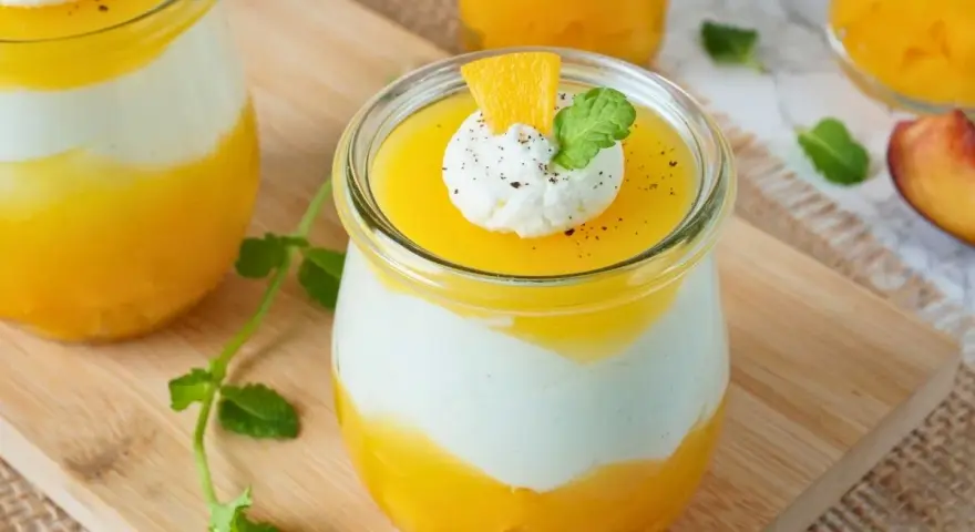 Erfrischendes Solero Dessert im Glas mit Mango-Fruchtsoße, weißer Creme und einem Klecks Sahne, garniert mit Minze und Orangenspalte.