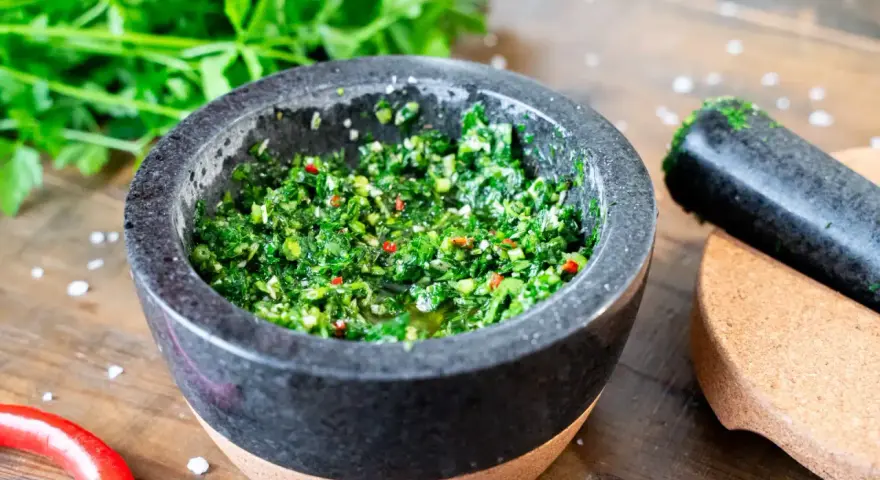 Frisches Chimichurri-Rezept im Mörser, bereit zum Verfeinern von Grillgut. Kräuter, Chili und Gewürze sind fein gehackt.