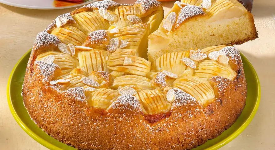 Ein saftiger, versunkener Apfelkuchen mit Mandelsplittern und Puderzucker bestreut, wird auf einem grünen Teller präsentiert.